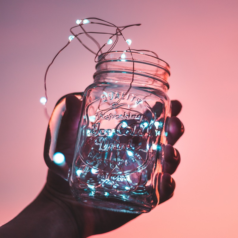 A hand holding up an embossed glass mason jar filled with tiny fairy lights, against a pink background