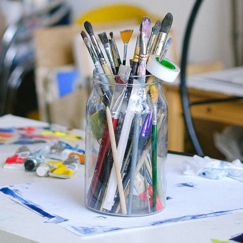 A mason jar holding paintbrushes, pencils and a roll of tape on a paint-splattered art table
