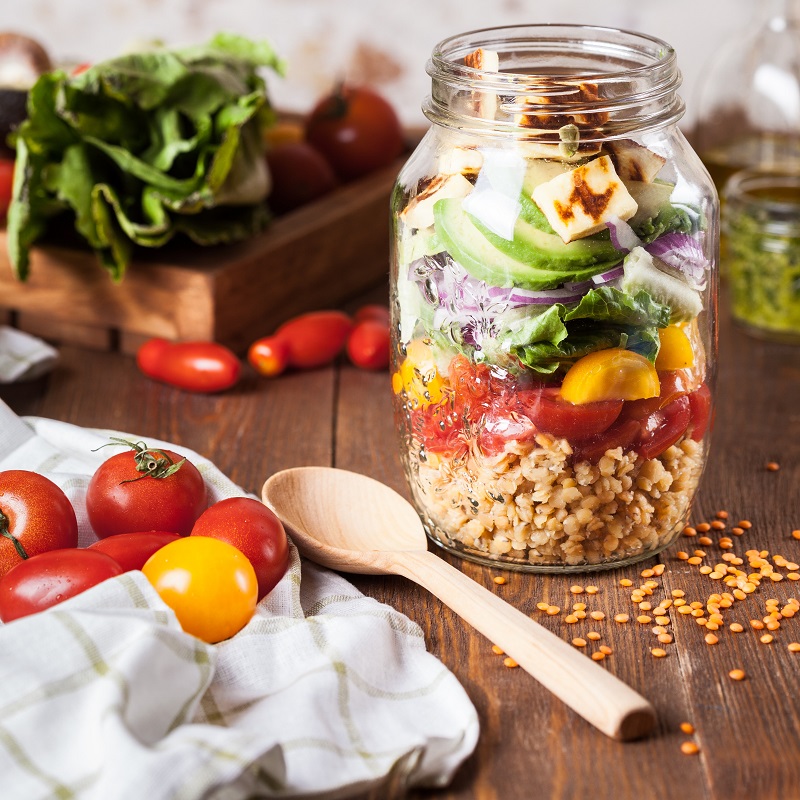 A layered salad in a mason jar of lentils, tomatoes, onion and greens, beside fresh tomatoes and a wooden spoon