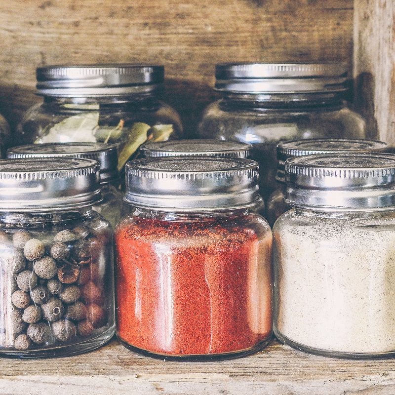 Whole and ground spices, including peppercorns and paprika, stored in glass jars with metal lids on a wooden shelf
