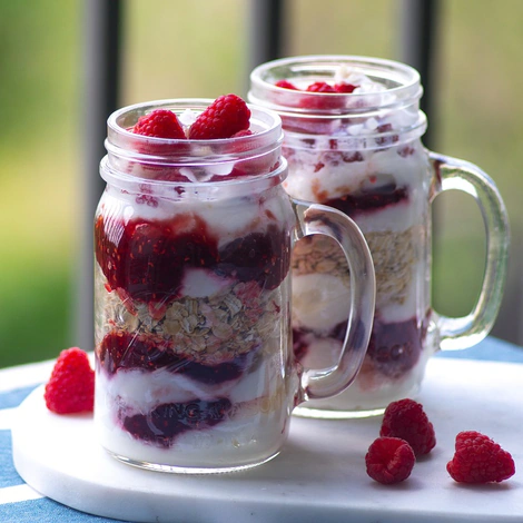 Two mason jar mugs layered with yogurt, granola and raspberries, with fresh raspberries on the side
