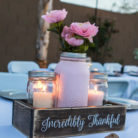 Table centerpiece: a white mason jar of pink roses between two candle jars, in a wooden box reading “Incredibly Thankful”
