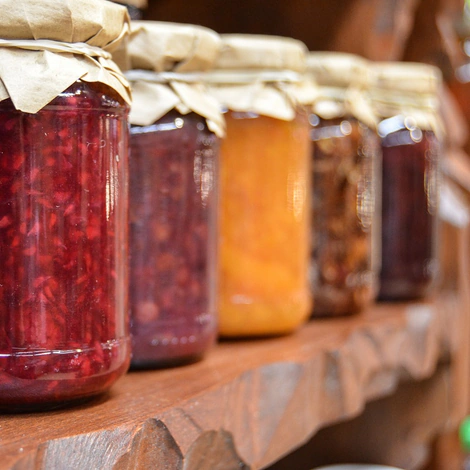 A row of homemade jams and preserves in jars with paper-covered lids, on a wooden shelf