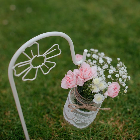 A lace-wrapped mason jar of pink roses and baby’s breath hanging from a white shepherd’s hook on a lawn, for a wedding ceremony
