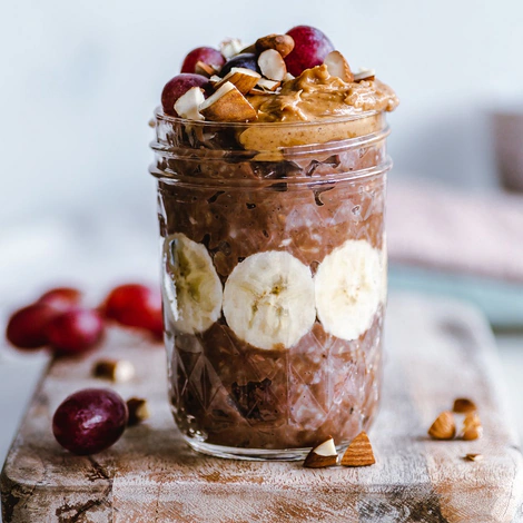 A mason jar snack layered with chocolate oats and banana slices, topped with peanut butter and nuts